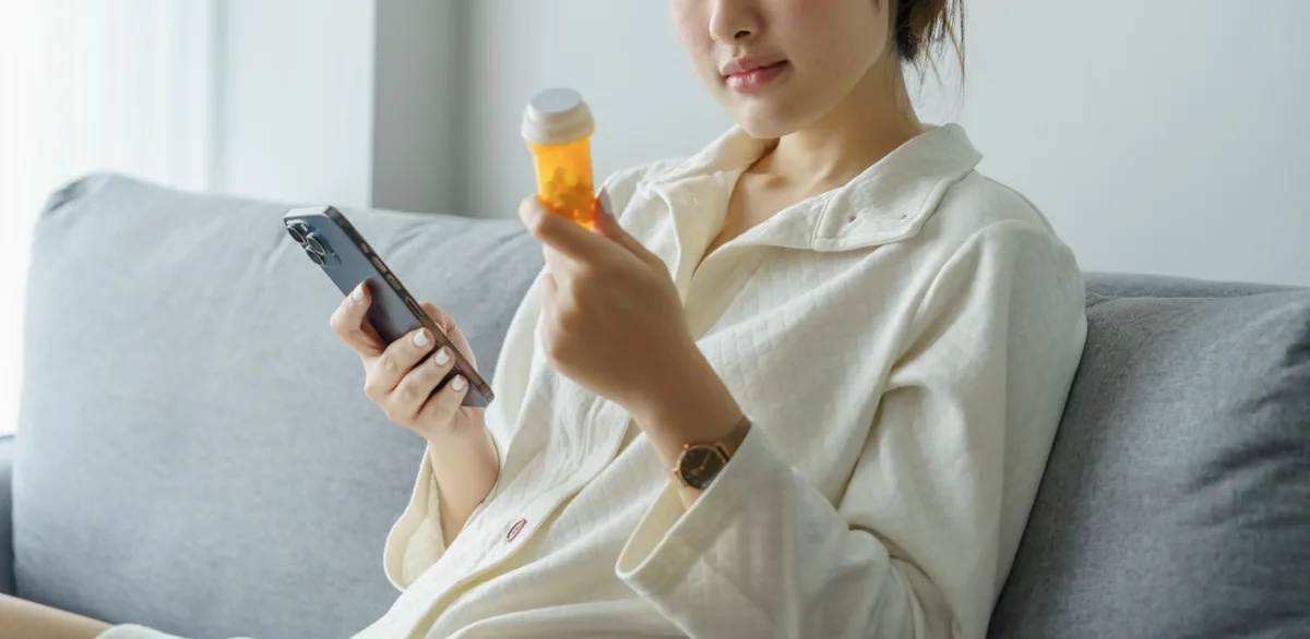 A woman reads a medicine label beside her phone at home.
